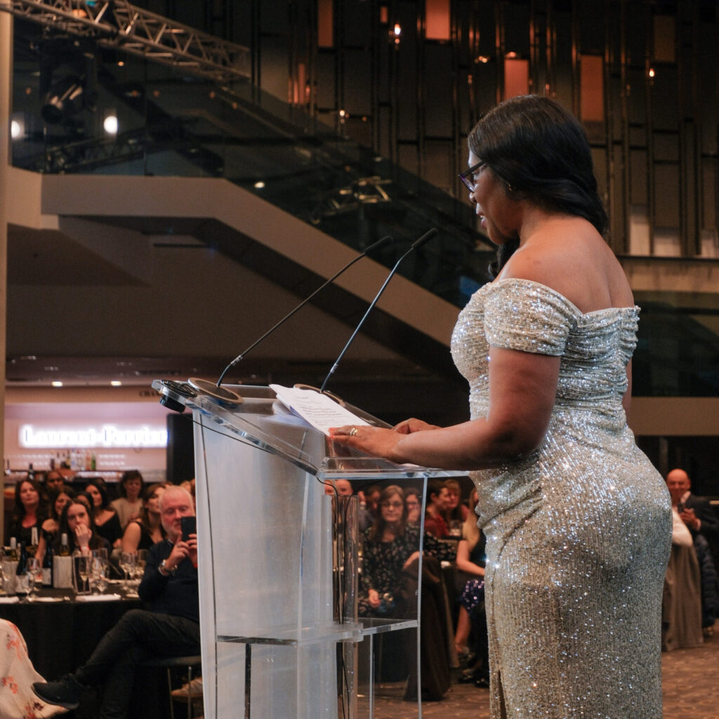 Audrey Bodman, a tall womain in a very sparkly silver dress, is at a lectern in front of an audience of people, presenting an award.