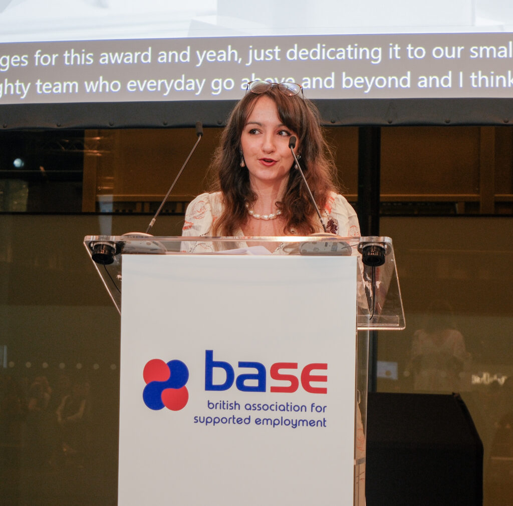 Beth Kume-Holland is stood at a lectern giving an acceptance speech for an award. The lectern has the BSAE logo on it, and in the background is a screen with live captions being shown.